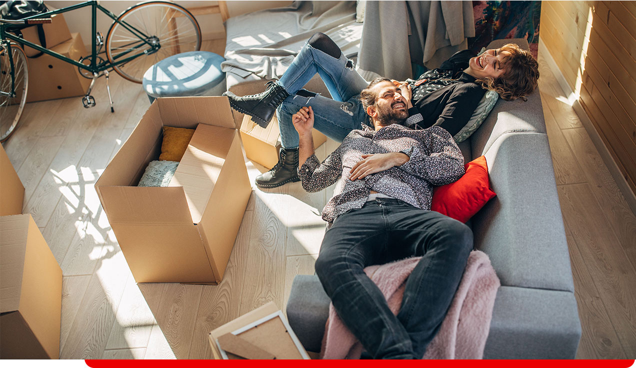 Zwei Personen liegen entspannt auf einem Sofa mit Umzugskartons und einem Fahrrad im Hintergrund